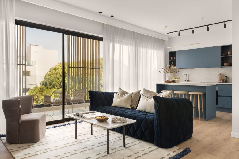 Chic and spacious living room in White City Limassol Aparthotel, featuring a deep blue velvet sofa, modern kitchenette, and floor-to-ceiling windows opening to a balcony.