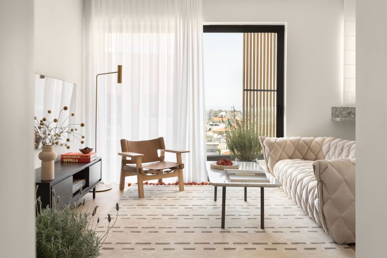 Chic living area with a plush beige sofa, contemporary wooden chair, and natural light flooding through sheer curtains and balcony doors.